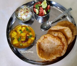 Khatti Meethi Aloo Sabzi | Potato Curry Recipe served on black platter with poori, shrikhand and salad on side