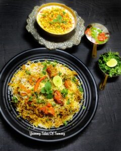 Chicken tikka biryani served in a black plate with boondi raita and salad on side