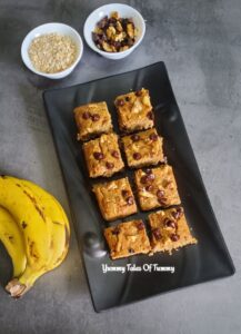 Sliced Air fryer Banana Oats Cake (eggless) served on a black tray with chocolate chips, Oats and banana on side